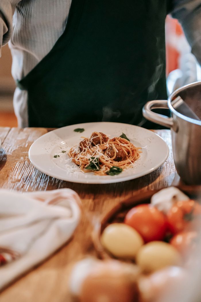 Crop unrecognizable man wearing apron while standing near counter and cooking pasta with meatballs and herbs on plate near tomatoes and garlic with lemons in light kitchen near pan